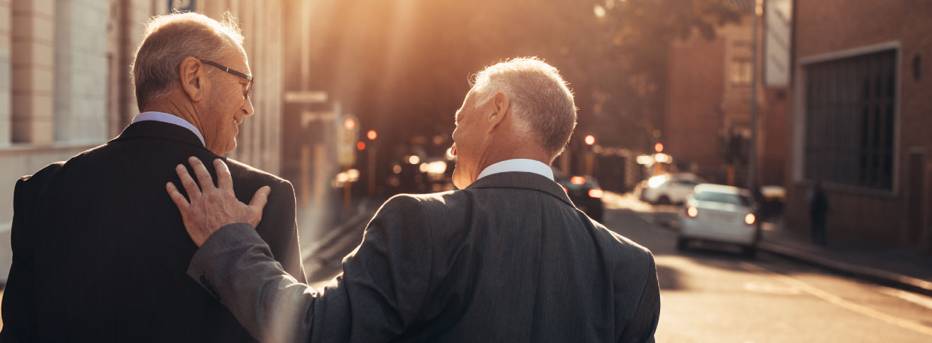 Two businessmen walking down a city street. One has his hand on the other's back. 