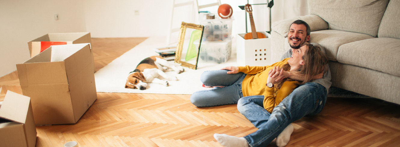 A young couple laughing and laying down in a half-empty room. There are cardboard boxes in the room.